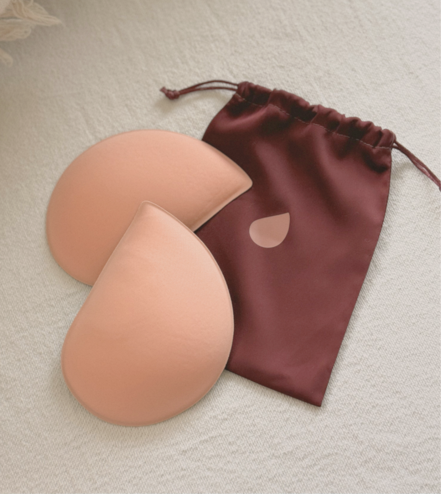 Two peach-colored, teardrop-shaped bra inserts lying next to a maroon drawstring pouch on a cream-colored fabric.