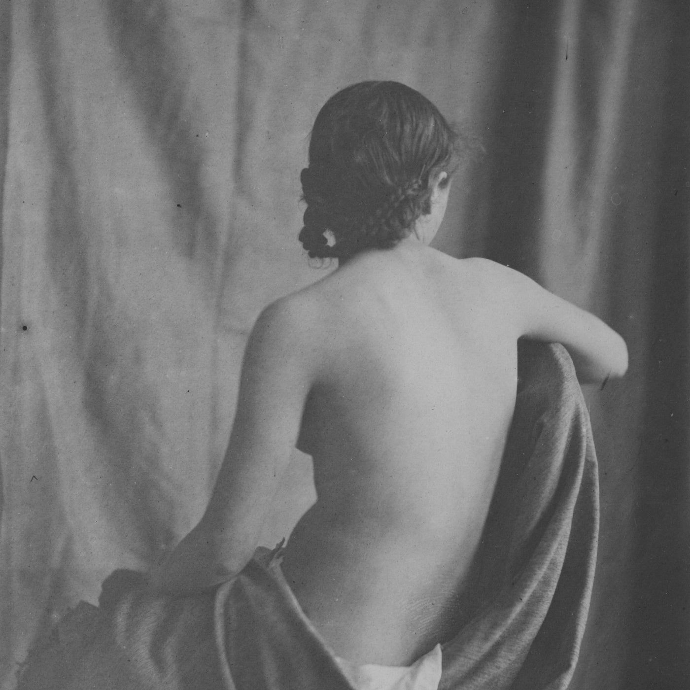 Black and white retro photograph of a nude woman draped with fabric against a plain background. We can only see her back. 